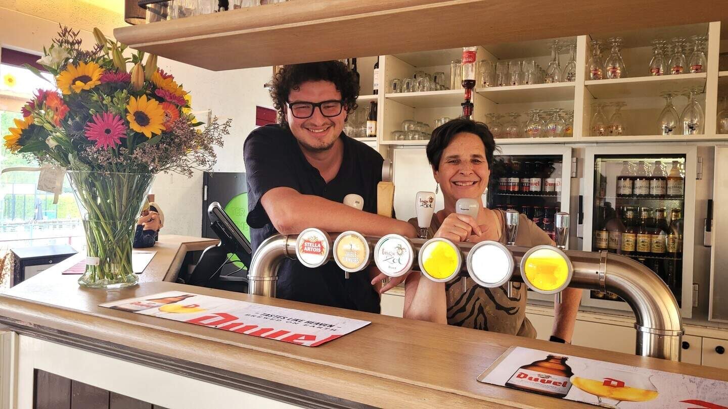 Jens en Veronique staan lachend achter de lichte houten toog van het café, met een grote bloemenbos en biertapkranen ernaast.